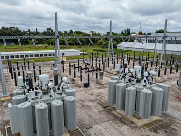 Wide panoramic overhead view of electrical utility substation switching yard, cool overcast natural light, transformer banks and high-voltage bus bars in tight geometric rows, sensor installation hardware visible on equipment housings, no people
