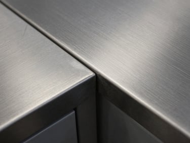 Extreme close-up overhead shot of a brushed inoxidable countertop edge joint, two panels meeting with near-invisible seam under cold task lighting that picks out the grain direction of each surface