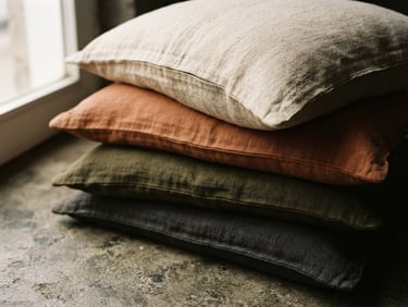 Close-up of heavy-weight linen pillow covers stacked on a concrete floor, soft morning window light, deep shadows, shallow depth of field.