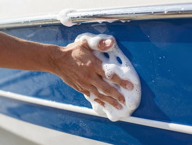 Close-up of a detailer's hand gently washing a boat hull with foam, bright morning light, water droplets, sharp focus