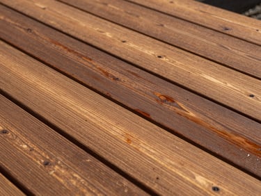 Outdoor timber decking being treated with a rich protective stain under bright Western Australian sunlight, showing wood grain.