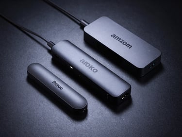 Flat-lay arrangement of a Roku stick, Amazon Fire Stick, and a small Android TV Box on a dark surface, cool backlight glow, shot from slightly above at a 30-degree angle