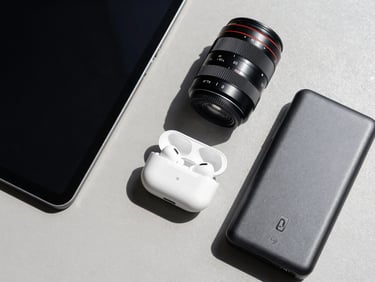 Flat-lay overhead shot of consumer electronics — a tablet, wireless earbuds case, and power bank arranged on a neutral gray surface, bright north-facing daylight, no branded packaging, product condition clear