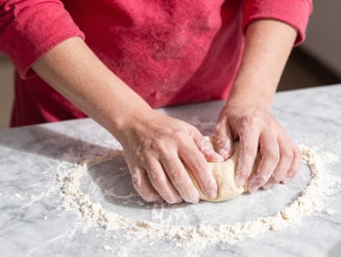 Close-up of hands in a vibrant red sleeve kneading artisanal dough on a flour-dusted marble counter, bright high-energy sunlight, high contrast.