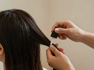Close-up of a stylist's hands applying a hair treatment product to dark strands on a salon chair, soft diffused natural light, warm skin tones against nude background, no face visible, precise and calm editorial framing