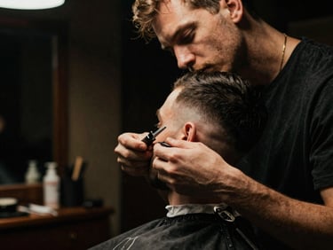 Moody cinematic photography of a master barber at work, dramatic low-key lighting, deep shadows, rich warm tones, shallow depth of field, 35mm. Focused shot of razor detailing a clean fade, warm interior glow.