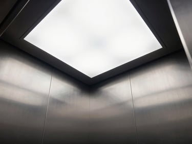 Low-angle interior shot of a premium elevator cabin, brushed steel panels, minimalist LED ceiling grid, sharp reflections, clean architectural lines