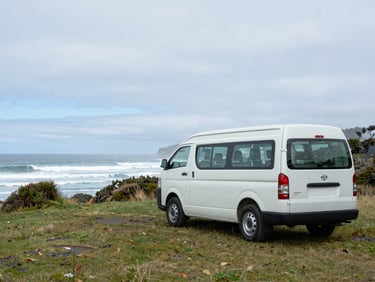 A self-contained Toyota Hiace campervan parked under an overcast sky at a coastal DOC campsite in Catlins, misty ocean waves in the background.