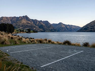 A private lakeside gravel parking spot in Wanaka, wide-angle lens showing the scale of the lake and mountains at dusk.