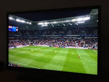 Tight framing on a live football match broadcast monitor, green pitch under floodlights, scoreboard overlay visible, high-contrast stadium lighting