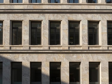 After: same commercial building facade now finished with printed monocapa render in morning light, impressed stone pattern visible in relief, strong shadow detail across surface