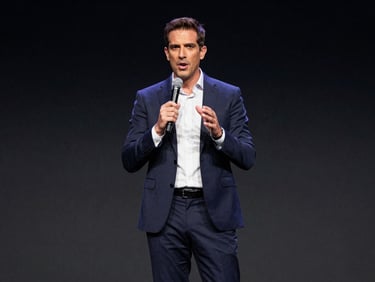 Medium-wide shot of Leo Colella commanding a corporate stage with a microphone, high-contrast dramatic stage lighting, deep charcoal background, professional executive posture, 35mm lens