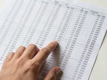 Overhead shot of a hand pointing at a printed analysis sheet covered in match statistics, confidence ratings, and circled odds — bright daylight, high contrast, no faces, clean desk surface visible at edges