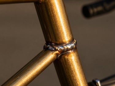 Extreme close-up of a polished brass fillet weld on a bicycle frame, deep shadows, warm editorial photography, 35mm