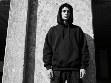 Low-angle heroic shot of a man in an oversized black hoodie standing against a raw concrete pillar, high-contrast monochrome lighting, sharp shadows, gritty editorial fashion photography.