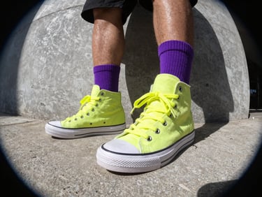 Dynamic low-angle fit pic on concrete featuring mismatched high-top sneakers, vivid purple socks, and neon lime yellow laces, harsh direct sunlight, raw urban aesthetic.