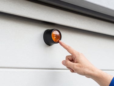 Close-up of a garage door safety sensor, with a technician's hand pointing to it, bright natural light.