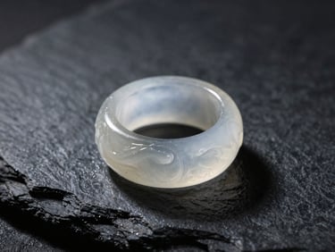 Macro photograph of a translucent white ice-jade sculptural ring resting on a dark slate surface, soft directional sunlight highlighting its internal water