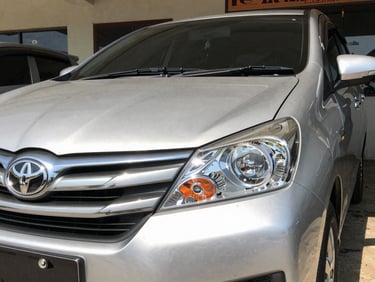 Close up of a clean silver Toyota Avanza front grille and sparkling clean headlight, parked outside a clean local showroom under direct bright Kupang sun, sharp focus