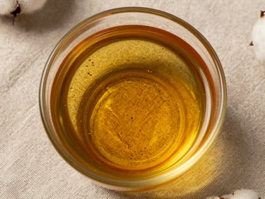 Extreme close-up overhead flat-lay of raw pressed cottonseed oil in a clear glass vessel on rough linen cloth, showing amber liquid clarity and oil surface texture, studio strobe from above, evidential framing