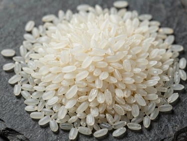 Extreme close-up overhead flat-lay of raw white rice grains on a dark stone surface, showing grain surface texture and translucency, documentary daylight from above, no lifestyle props