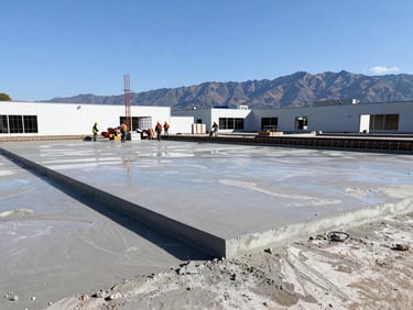 Wide angle of a commercial building foundation pour, wet concrete, active crew in safety gear, crisp daylight.