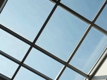 Low-angle shot looking up through a structural glass skylight framing a clear blue sky, geometric steel support lines, bright diffused daylight, 35mm