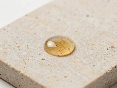 Macro texture shot of a thick, translucent amber serum droplet on a warm travertine stone slab, soft natural daylight, minimalist laboratory setting