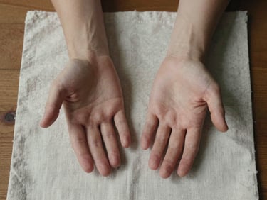 Overhead view of two hands resting open on a linen cloth, soft overcast daylight, wooden table beneath, calm and still