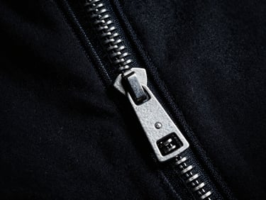 Macro photography of matte silver zipper detail on deep black heavy cotton fabric, dramatic high-contrast side-lighting, sharp metallic highlights, medium format film.