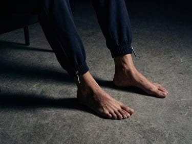 Moody editorial crop of tailored jogger cuffs resting against minimalist concrete floor, sharp highlights on matte silver drawcord tips, deep shadows.