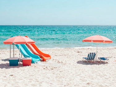 Vibrant beach setup with a water slide, colorful coolers, and umbrellas on a white sand beach under harsh midday sun, sparkling gulf water