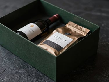 Close-up of a modern, minimalist gift box, featuring a bottle of high-end wine and a bag of specialty coffee, on a dark stone surface, low-key lighting, rich shadows.