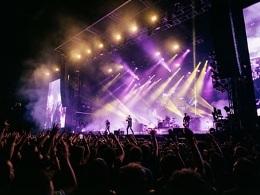 Low-angle wide shot of a massive concert stage, dramatic purple and gold theatrical lighting beams cutting through deep shadows, haze in the air, crowd silhouettes in the foreground, 35mm film style.