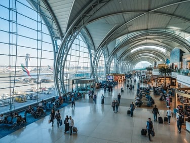 Wide environmental shot of a modern, sunlit airport terminal in Dubai, architectural steel structures, airplanes visible through glass walls, cool blue and gold tones, 35mm.