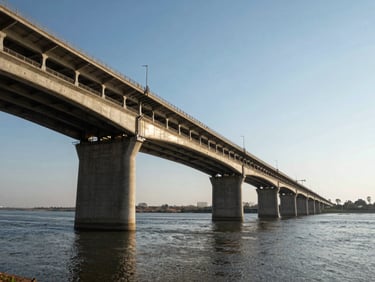 Completed multi-span concrete highway bridge stretching across a wide river, harsh midday sun reflecting off the structural steel piers, wide-angle lens