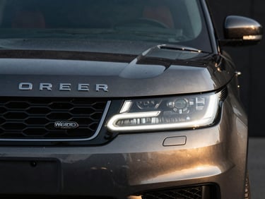 Front grille detail of a dark Range Rover Sport, polished black paint reflecting subtle golden light, dark background