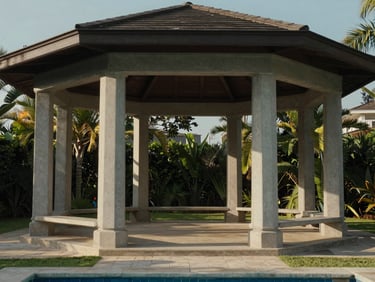 Eye-level view of the Royal Pool Villa gazebo structure from inside the plot — solid roof, clean concrete pillars, afternoon light casting defined shadows on the paved floor beneath, garden visible beyond