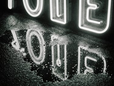 High-contrast monochrome close-up of neon signs reflecting in a rain puddle on asphalt, raw film grain, green tint