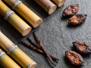 Overhead flatlay of raw ingredients, unrefined sugar cane, vanilla pods, and cacao beans on a dark stone surface, warm side-light, 35mm