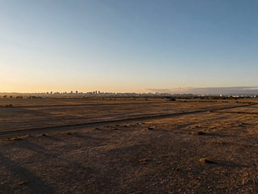 Wide panoramic shot of undeveloped land in Peru's coastal growth corridor at golden hour, flat terrain extending to the horizon, distant city outline visible, long shadows across the earth