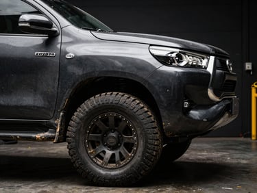 Dramatic close-up of a customized Toyota Hilux pickup truck, dirty tires with deep treads, high-contrast dark garage background, sharp metallic details.