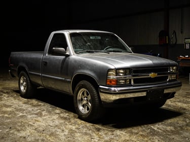 Low-key high-contrast photo of a modified Chevrolet S10 truck, dark silver metallic paint reflecting sharp overhead workshop lights, gritty concrete floor.