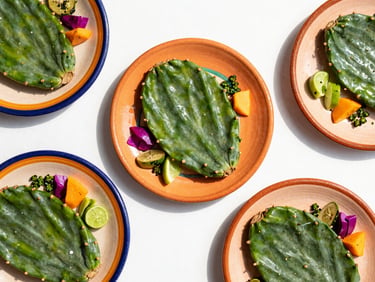 Top-down flat lay of vibrant Mexican plates with fresh green nopales, colorful garnishes, and bright daylight.