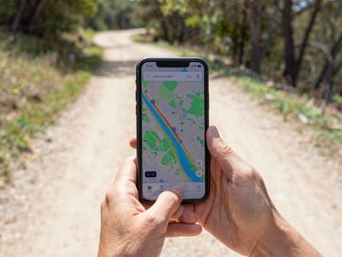 First-person perspective of a hand holding an Android phone displaying a colorful map interface on a sunny dirt hiking trail, trees blurred in background, 35mm lens.