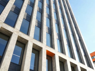 Wide-angle architectural shot of a modern concrete and steel skyscraper facade under bright, high-key natural daylight, casting sharp geometric shadows with electric blue sky reflections.