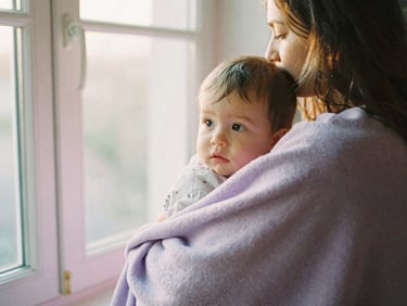 A mother cradling her child in a soft lilac blanket, looking out of a sunlit window, gentle warm light, 35mm film.