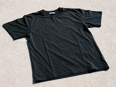 Detail shot of a heavy black cotton t-shirt with reinforced seams, laying flat on raw light gray concrete, harsh sunlight