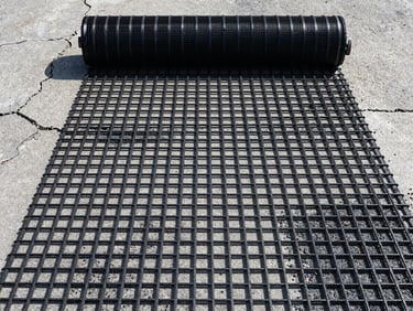Top-down flat-lay shot of black fiberglass geogrid mesh being unrolled over an old, cracked concrete pavement before asphalt overlay, emphasizing the grid pattern, harsh direct sunlight, sharp focus.