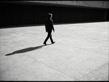 An isolated figure walking across a vast empty brutalist concrete plaza, harsh direct sunlight, long deep shadow, 35mm film